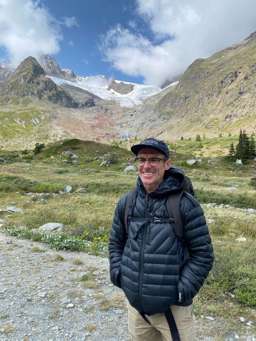 Aaron Winning smiling on a mountain trail, with a glacier behind him